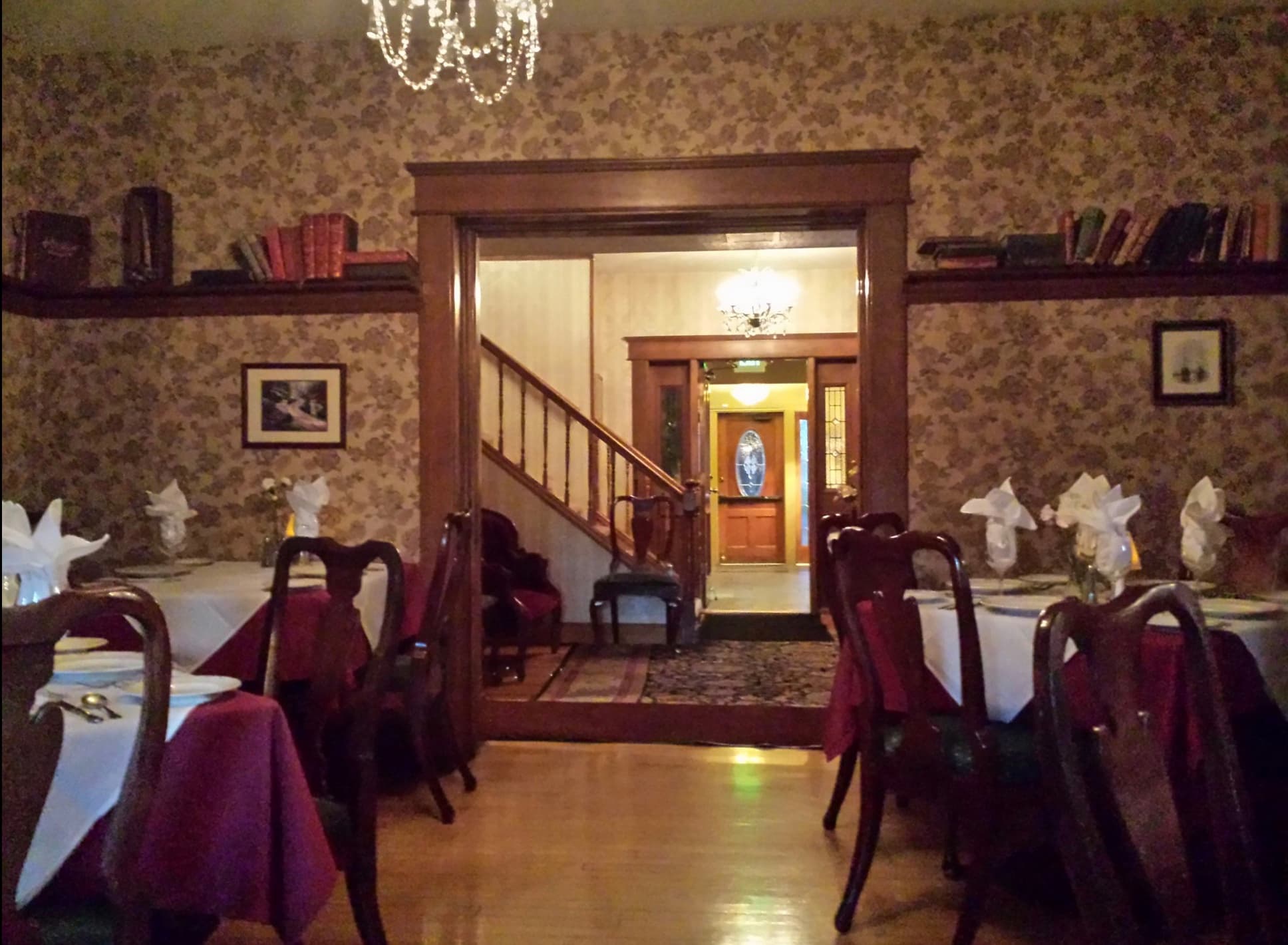 A Victorian dining room inside the 1908 Historic House, with a chandelier, papered walls, and the foyer staircase visible through the doorway